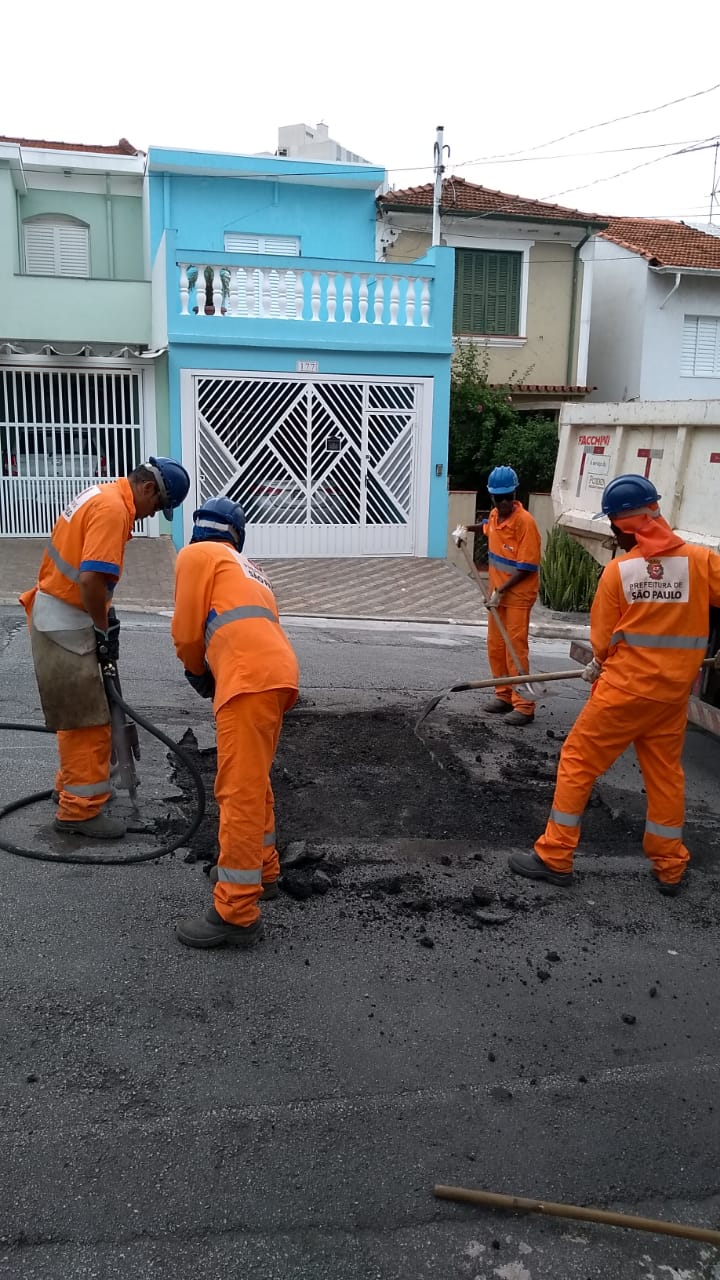 Foto: Quatro funcionários da Subprefeitura Ipiranga, vestidos com uniformes na cor laranja e capacetes na cor azul, realizando o serviço de tapa-buraco na rua Frei Durão
